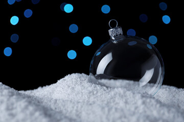 Transparent Christmas ball on snow against blurred fairy lights, space for text