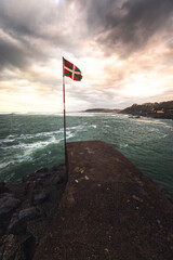 Fototapeta premium Basque national flag (Ikurriña) at the edge of a pier on the coast; Getaria, North Basque Country.