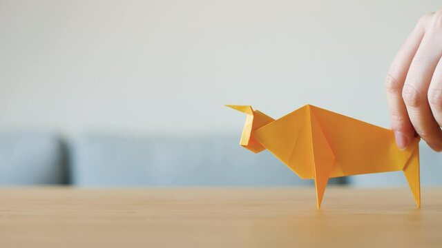 A hand moving yellow Origami Ox on table as symbol for a happy Chinese new year. Cow or bull is the Chinese zodiac sign for 2021. Animal made with traditional Japanese paper folding technique. 4K.