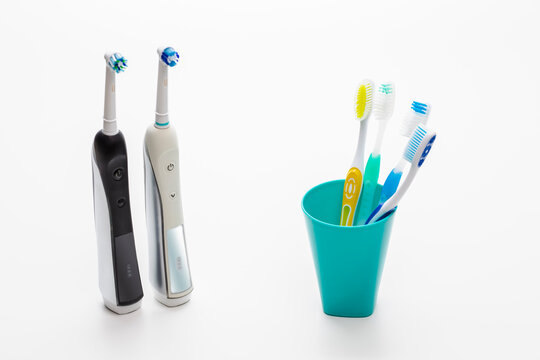 Pair of Professional Electric Toothbrushes In Front of Four Manual Tooth Brushes in One Cup On White Background.