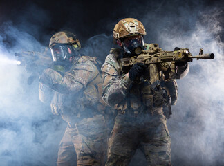Two soldiers in military gear, bulletproof vests and gas masks cover each other and raise their submachine guns taking aim, in full combat readiness to break through the smoke from chemical weapons