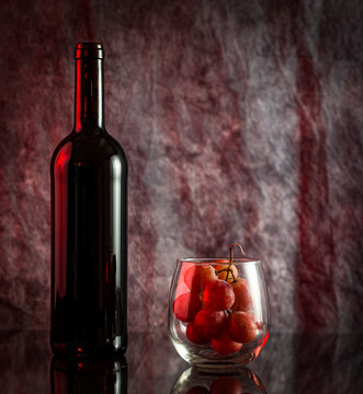 Bottle Of Red Wine With Fresh Grape Inside Stemless Wine Glass On A Reflective Surface. High Definition Picture. Added Post-crop Vignetting For A Moody And More Intimate Effect.