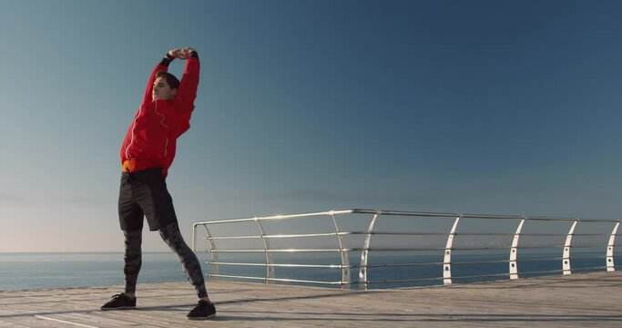 Young handsome guy in sports uniform doing physical exercises standing on outside against the background of the sea and sunlight.