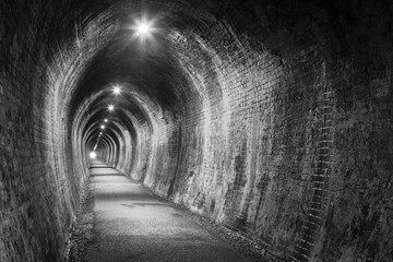 An old tunnel with brick walls, lit up with electric lighting. Black and white. Photographed at the...