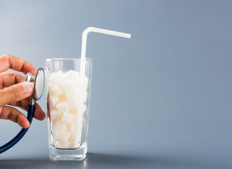 Hand of doctor hold stethoscope check on glass full of white sugar cube sweet food ingredient, isolated on gray background, health high blood risk of diabetes and calorie intake unhealthy drink