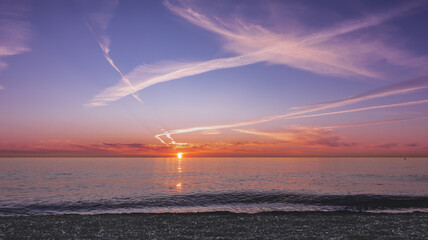 Sunset over the sea. The sun is low on the horizon. The sky is highlighted in orange, you can see the traces of flying planes. There is a sunny path on the water. A wave rolls on the pebble beach.