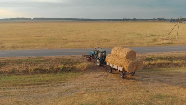 Aerial view of a farmer on a tractor with a full trailer of bales of straw or hay, vyzzhayuschim from the field to the paved road in the countryside in Sunny evening weather. Stock of animal feed