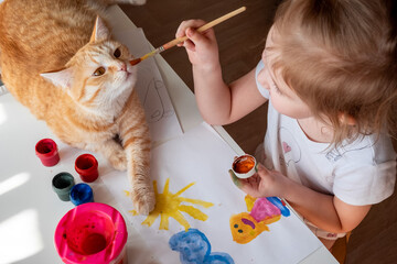 A little girl paints the sun and her mother with watercolors, a ginger cat lies next to the table. Children's creativity. Distance learning, top view