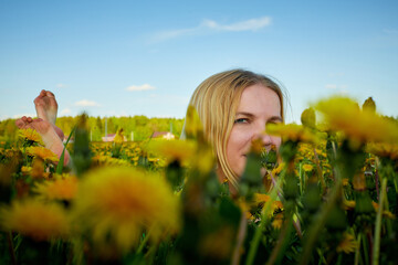 Pretty woman on a field with green grass and yellow dandelion flowers in a sunny day. Girl on nature with yellow flowers and blue sky.