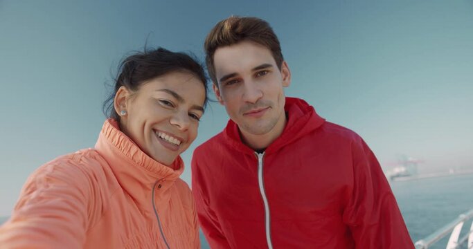 Young happy couple recording video message standing on background of the sea in tracksuits after morning jog. View from camera.