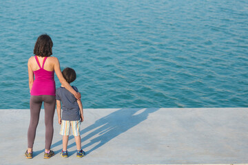 A boy with his mother stand on the promenade and look at the water.