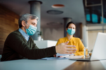 Businessman and his female colleague wearing face masks while working on laptop in the office.