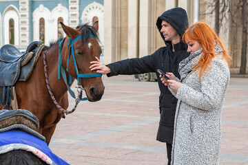 Young man and red-haired woman stand on the street of the city. A man strokes a horse's head with...