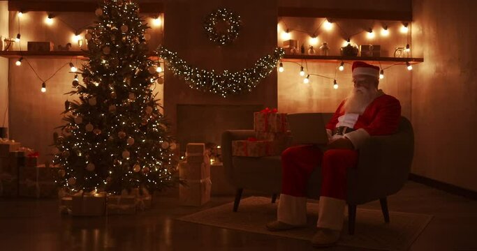 Santa Claus is working on a laptop sitting at home on the sofa in the background of Christmas. Santa responds to children emails