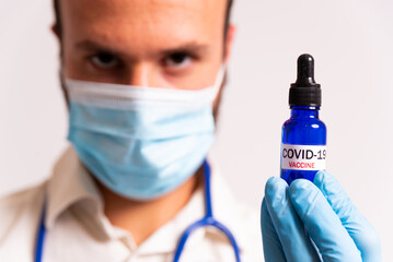 young male doctor holding covid-19 vaccine blue vial. concept of science and corona virus vaccine. face mask and stethoscope on white background.