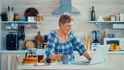 Senior woman drinking coffee and waving during a video conference with family in kitchen while having breakfast. Elderly person using internet online chat technology video webcam making a video call