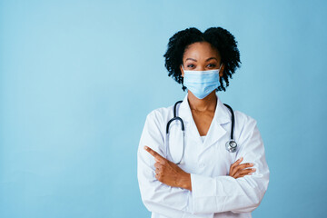 Portrait of a doctor in lab coat with face mask and arms crossed pointing to side at copy space