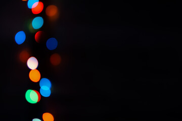 Multi-colored bokeh on a black background. Bright blurry textures of holiday lights. Colorful beautiful Christmas garlands on blurry background. Layout of a greeting card for the New year. Copy space
