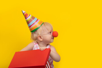 Funny little girl dressed as clown opens gift box and looking to side. Portrait on yellow background
