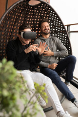 Amused gay couple testing the virtual reality glasses at home. Sitting in the swing on the balcony. High quality photo