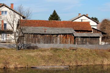 Elongated old wooden barn with storage area and wooden corn shed built next to calm river protected with flood protection wall made of box barriers covered with thick dark grey geotextile fabric