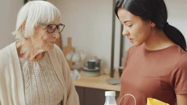 Close up view of young Asian female volunteer talking with senior Caucasian woman while unloading groceries from paper bag for her on kitchen table at home