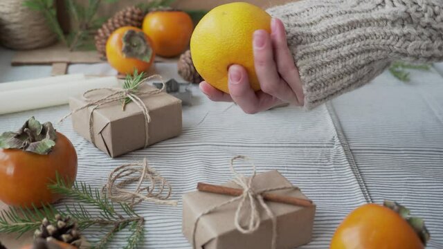 A girl in a knitted sweater holds a tangerine in her hands, surrounded by Christmas gifts. Christmas concept, new year's holiday atmosphere. Craft paper gift wrapping handmade.