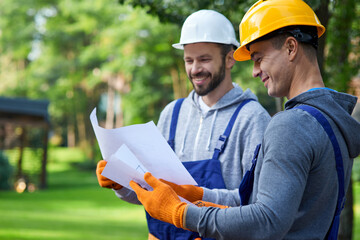 Discussing ideas. Portrait of two positive builders standing outdoors with an open blueprint discussing the half completed cottage house