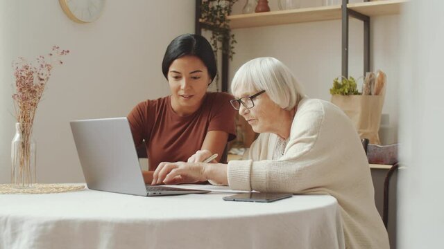 Young Asian female caregiver sitting at table with elderly Caucasian woman and teaching her how to use laptop at home