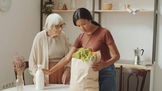 Young Asian female volunteer smiling and speaking with thankful elderly woman while unloading groceries from bag for her at home