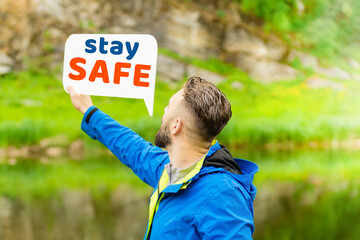 Naklejka premium A young man in a blue jacket stands in the nature in summer and holds the message on the banner stay safe. The concept of social distance in the fight against pandemic 2020. quarantine