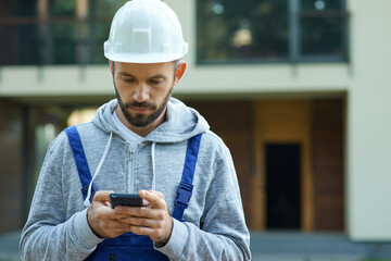 Young male builder in hardhat using smartphone, standing outdoors while working at cottage...