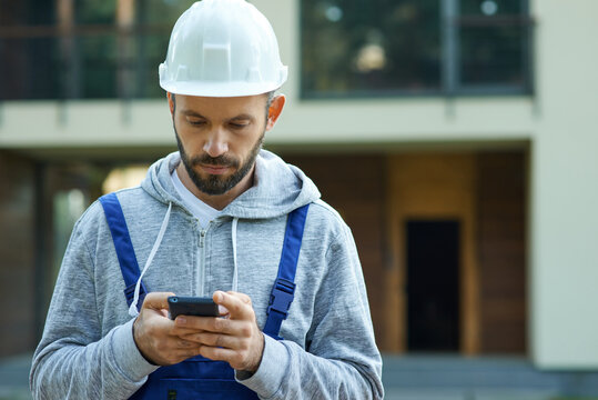 Young male builder in hardhat using smartphone, standing outdoors while working at cottage construction site