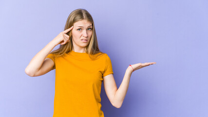 Young blonde woman isolated on purple background holding and showing a product on hand.