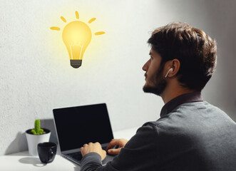 Portrait of young guy looking at yellow light bulb; having new idea. Using laptop and wireless earbuds or earphones.