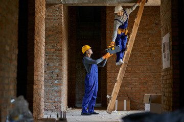 Assistance. Full length shot of young workman in blue overalls and hard hat giving toolbox to his...