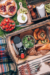 Great picnic basket with cheese, strawberries, grapes, baguette, pizza, salad, wine for picnic on plaid over green background. Top view. Copy space. Summer family lunch. Romantic picnic