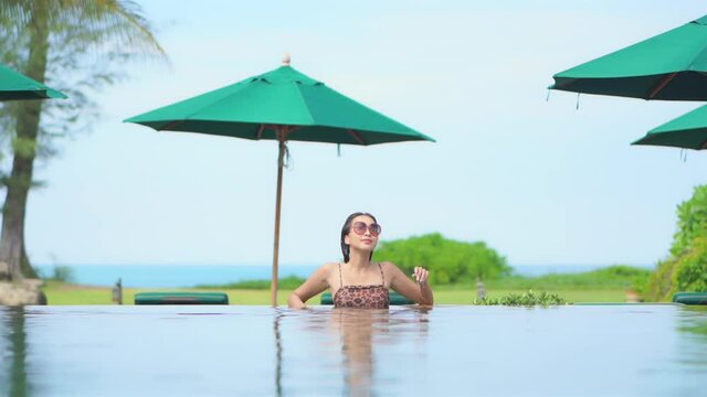 Asian model standing in swimming pool with sunglasses looking at tropical paradise landscape of island resort with ocean and green umbrellas in background.