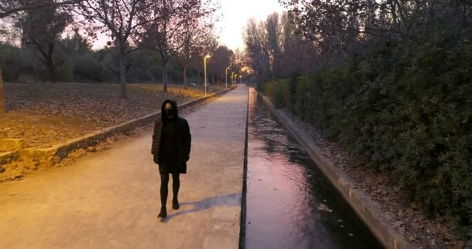 mujer de negro con capucha y mascarilla covit paseando en parque oto&ntilde;al y canal de agua