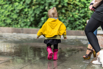 Two young mothers of the girl are watching a small child walking through puddles in a yellow raincoat and pink rubber boots. The concept of childhood. Universal Children's Day.