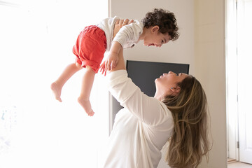 Happy mom holding excited baby in arms, lifting child in air. Side view. Parenthood and childhood concept