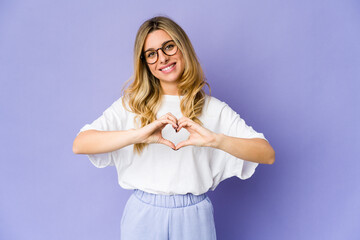 Young caucasian blonde woman smiling and showing a heart shape with hands.