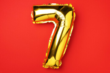 Creative layout. Golden foil balloon number and digit seven 7. Birthday greeting card. Anniversary concept. Top view. Copy space. Stylish gold numeral over red background. Numerical digit.