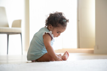 Pensive dark curly haired baby girl wearing pale blue dress, sitting on floor at home, touching...