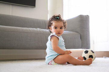 Sweet serious black haired baby girl in pale blue clothes sitting on floor with soccer ball,...