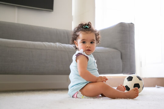 Sweet serious black haired baby girl in pale blue clothes sitting on floor with soccer ball, looking at camera. Side view. Kid at home and childhood concept