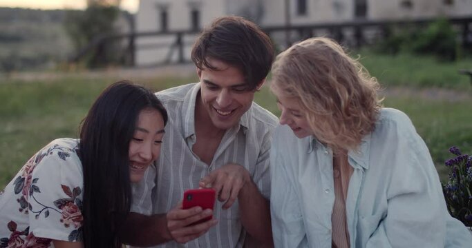 Young cheerful man and two women looking at phone screen and talking.Smiling diverse friends using smartphone and communicating while sitting on grass at park. Concept of friendship