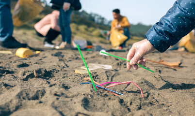 Woman hand picking up straws on the beach with group of volunteers working in the background. Selective focus in straws in foreground
