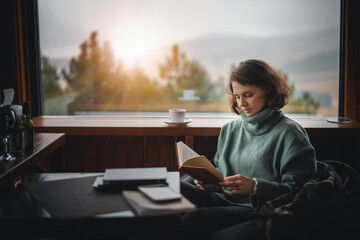 Young woman in green sweater reading a book and drinks coffee by the window in her country house with a view of the mountains