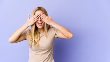 Young blonde caucasian woman covers eyes with hands, smiles broadly waiting for a surprise.